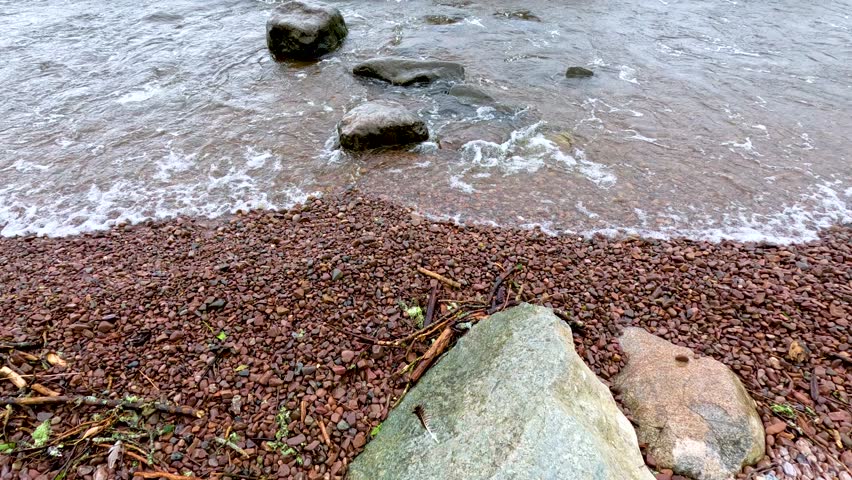 Overhead view of gentle waves washing over a rocky, pebbled shoreline at Loch Ness, Scotland.