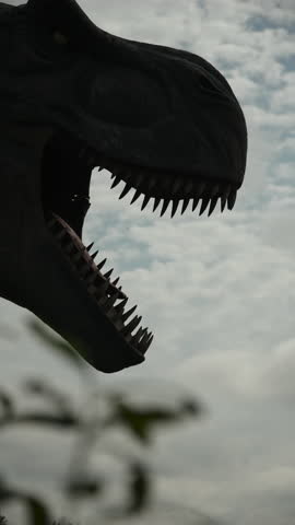 Silhouette of bobbing head with open mouth, teeth of predatory animal against cloudy sky. Vertical video of mechanical model of reptile near plants.