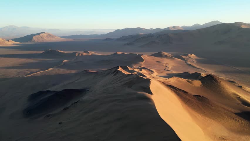 Aerial drone view of vast golden sand dunes glowing under the warm sunset in the Chilean Atacama Desert.