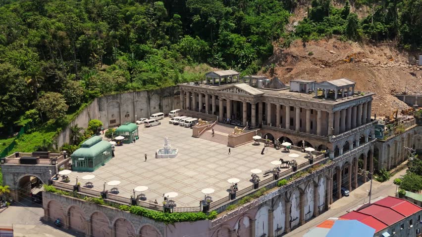 Drone shot of a grand neoclassical-style building nestled on a hilltop, framed by tropical forest and ongoing hillside development.