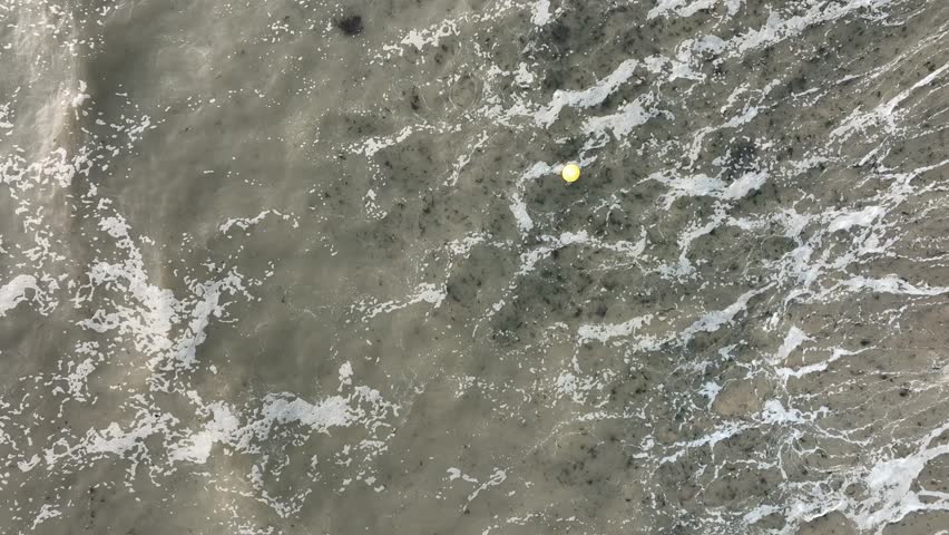 Aerial view of ocean water with foamy waves and a yellow ball floating on the surface, Bernières-sur-Mer, Normandy, France.