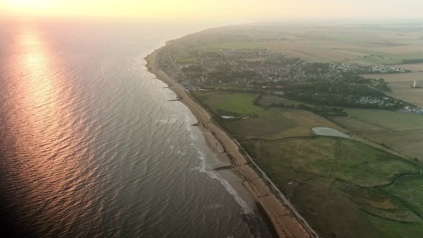 Aerial view of the sun reflecting on the ocean meeting the sandy beach and lush green fields near Bernières-sur-Mer, Normandy, France.