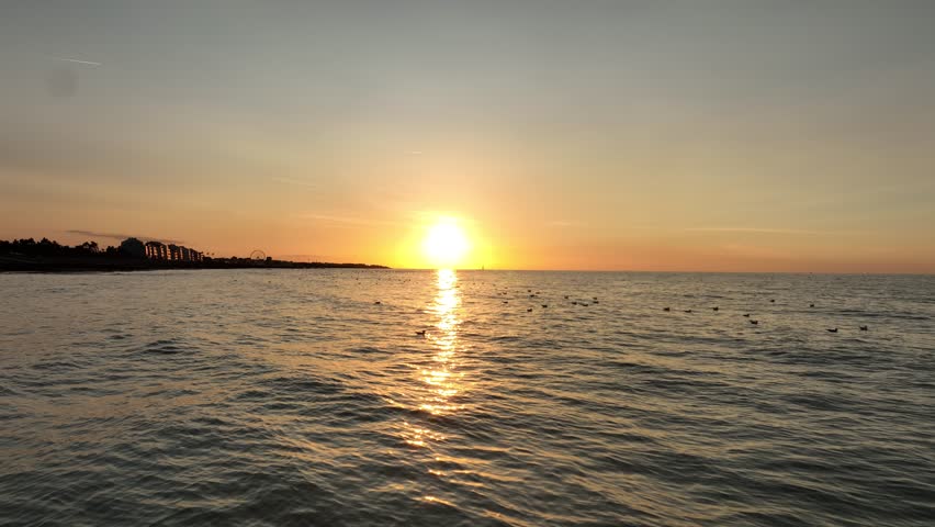 Aerial view of the sun setting over the sea with birds flying over the water in Bernières-sur-Mer, Normandy, France.