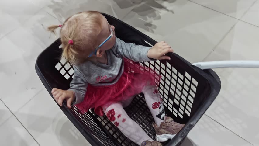 Small girl in glasses enjoying ride in shopping basket. Happy time shopping with parent. Children fashion. Cheerful carefree retail visit, chosen wardrobe, childhood innocence. Candid, joyful energy