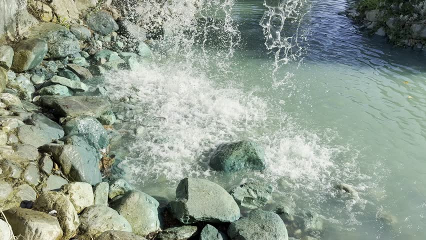 Cinematic closeup waterfall in landscaped summer park, stone banks and flowing stream, camera rising slowly along falling water capturing sparkling drops and spray, trees, gazebo, bridge in background