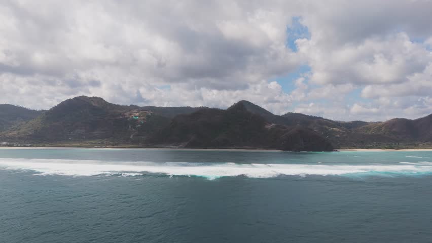 An expansive aerial view of the ocean near Lombok, Indonesia. Waves roll toward the shore, with lush green mountains in the background, creating a stunning coastal landscape