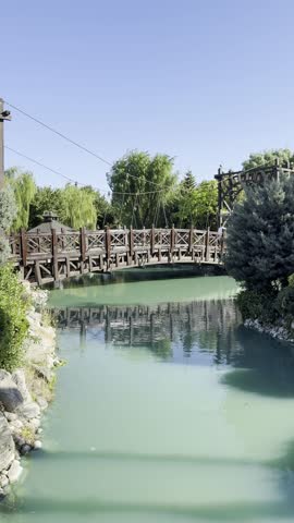Stylish Wooden Bridge over Small Stream in Urban Park with Stone Banks, Green Trees, Bushes and Reflections in Calm Water under Bright Blue Summer Sky. Scenic Landscape with Relaxing Nature Sounds.