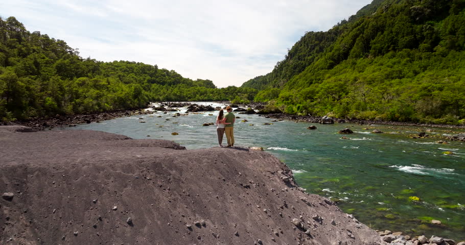 Couple standing on volcanic sandbank, admiring scenic Petrohué River flows through lush green forest, Chile. Aerial forward overhead