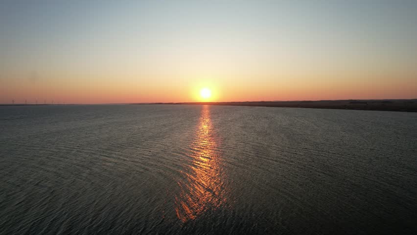 Aerial view of a beautiful sunset over the sea with orange sunlight reflecting on the water surface. Scenic horizon with calm ocean waves and wind turbines in the distance