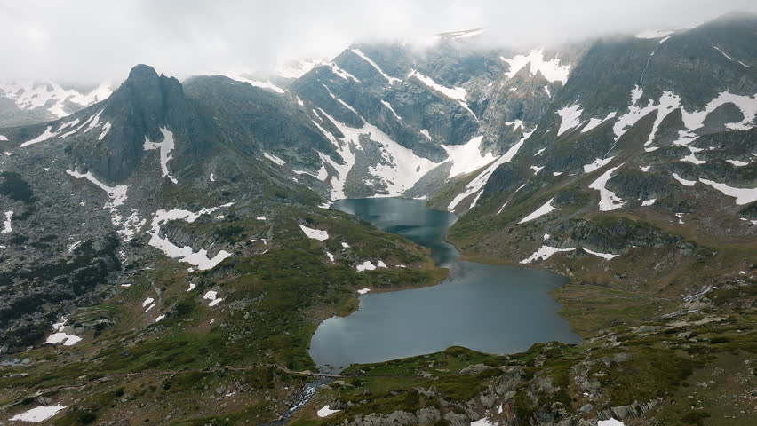 Alpine wilderness captured from above in Seven Rila Lakes Bulgaria, featuring crystal lake, fog, summer snow, and rocky slopes, great for adventure, drone footage, nature stock buyers, and scenic