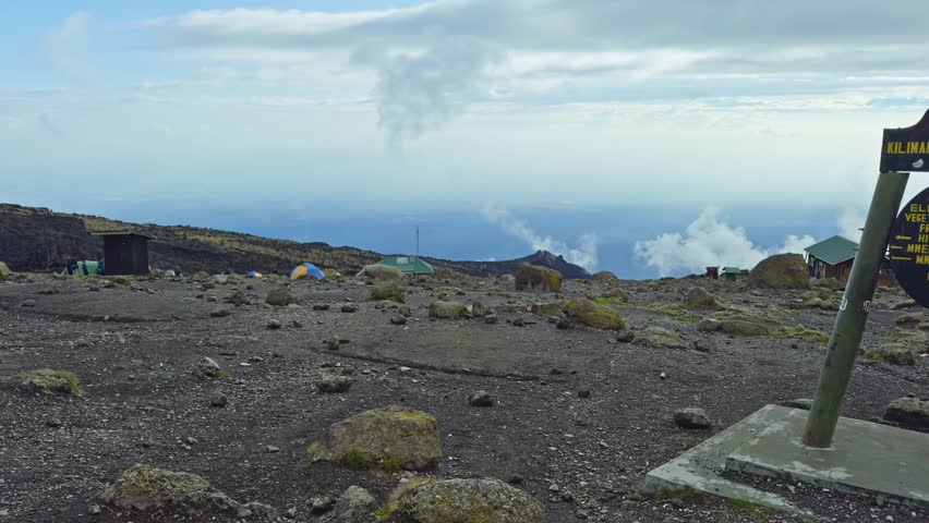 Ground-level pan reveals a high-altitude volcanic plateau with a Kilimanjaro National Park sign on a concrete base, surrounded by scattered rocks, small huts and clouds.