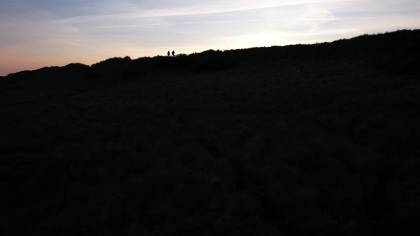 Aerial view of silhouettes of two people standing on coastal dunes during a colorful sunset. Scenic evening sky with orange and purple tones over the sea horizon.