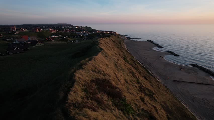 Aerial view of a coastal landscape with sandy beach, grassy dunes and calm sea during sunset. Scenic shoreline with houses on the hillside and clear evening sky in Denmark.