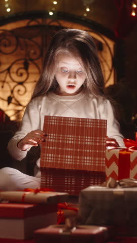 Little girl opening Christmas gift box with magical light by the fireplace