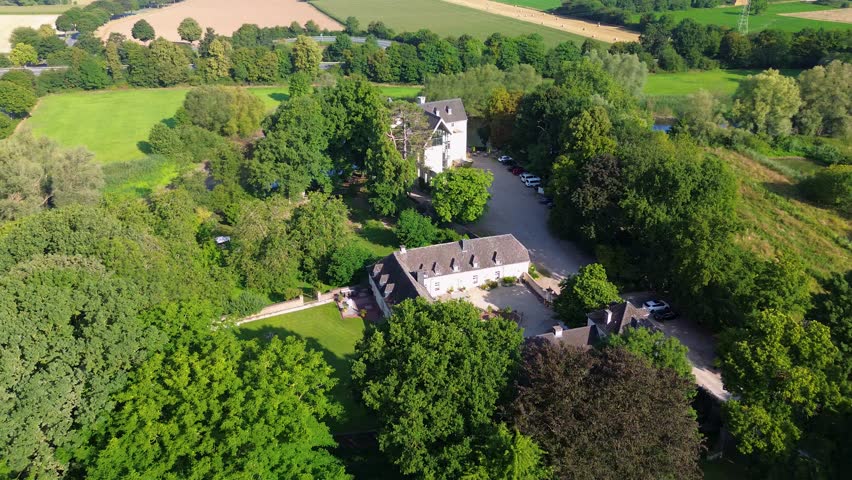 A charming historic estate with a U-shaped courtyard, surrounded by lush greenery, fields, and trees, blending architecture with serene natural beauty.