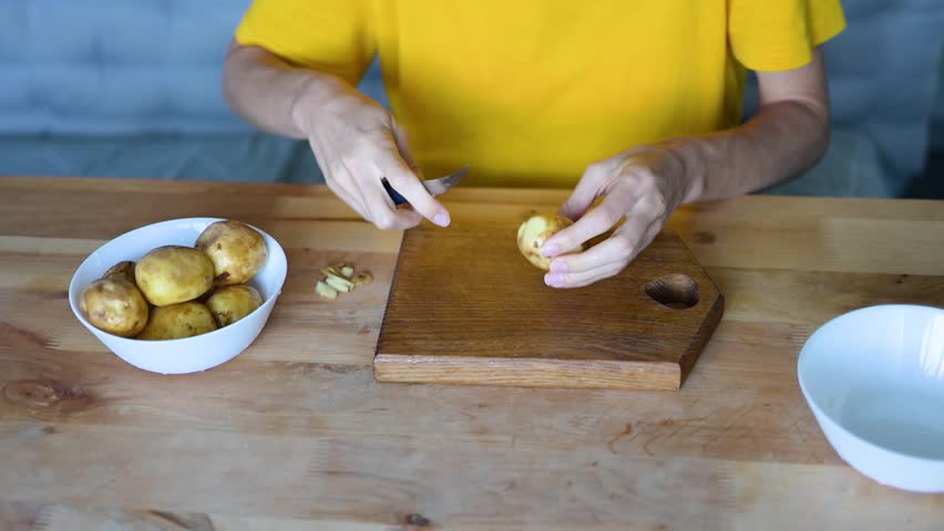 Hands peeling a potato over a wooden cutting board, with a bowl of potatoes an empty bowl nearby, ready for preparation fullhd video food