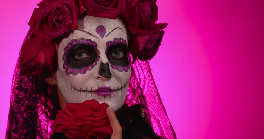 A girl with elaborate calavera catalina makeup, adorned with flowers, poses against a bright pink background. She is celebrating the Day of the Dead.