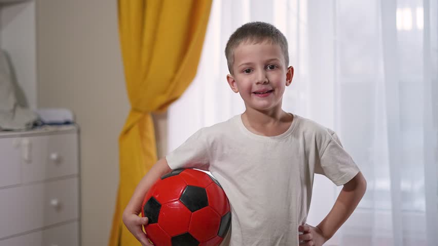 Playing boy holds football ball confidently. Young child enjoys sport activity with bright soccer ball. Sport develops young character. Football game for active child lifestyle indoors sport game.