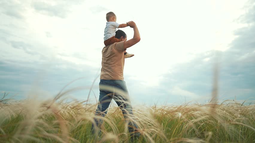 Father bonding with child walking across grass. Man holds son walking slowly. Boy smiles during bonding moment. Parent walks with son. Child walking on field with father. Family bonding outside.