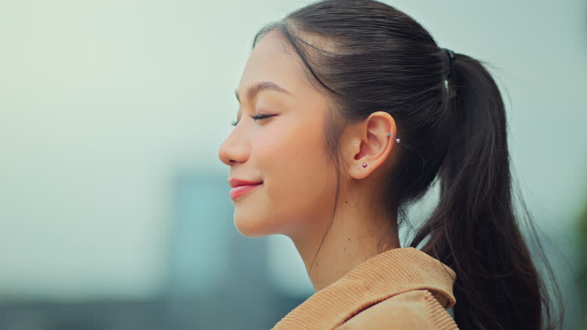 Cheerful Asian woman closing eyes and breathing deeply while enjoying fresh air in urban park, relaxing outdoors with mindful wellness lifestyle