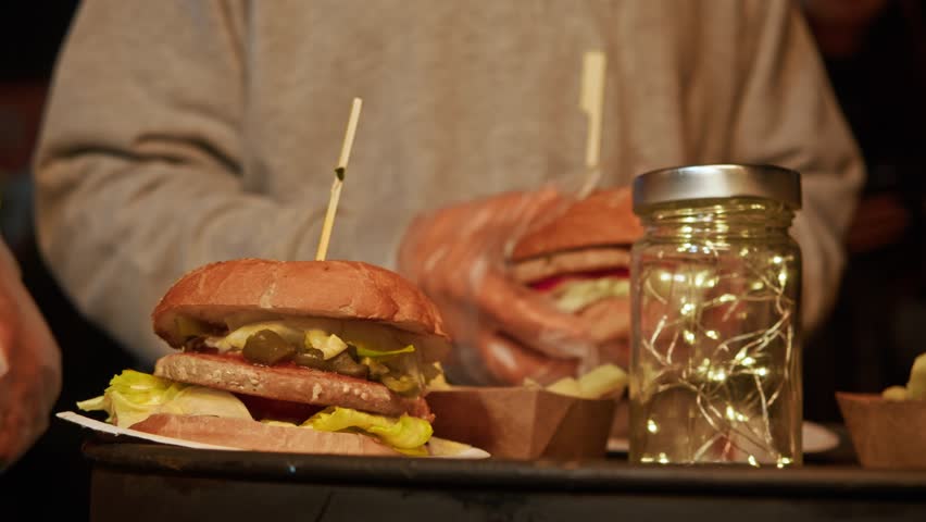 Two people wearing transparent gloves, at a nightclub outside, start eating juicy, mouth-watering burgers. Close up