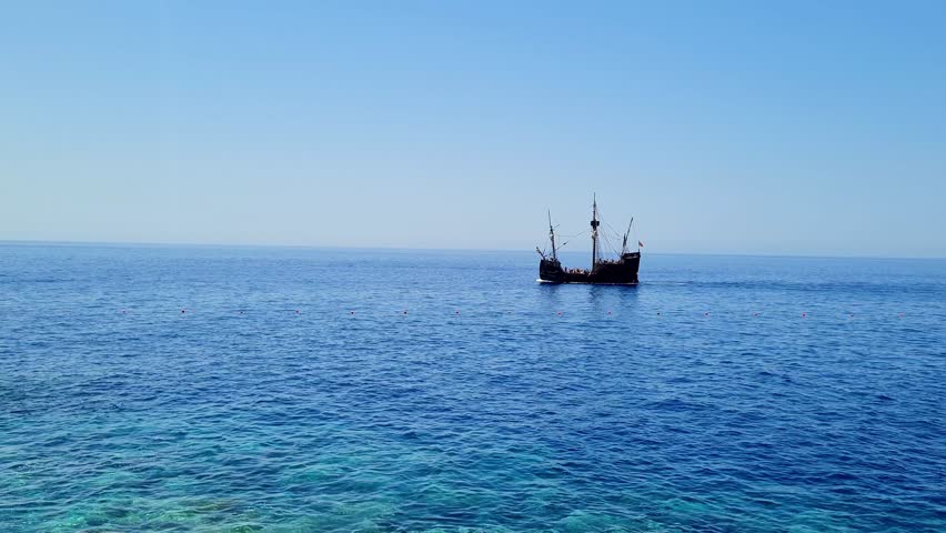 An old wooden ship sails on the ocean.The sky is cloudless and merges with the horizon line.The water has several shades of blue - from light turquoise in the foreground to dark blue in the distance