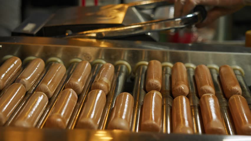 Roller grill with sausage filling, turning over under constant heat while the cook roasts a hot dog bun on the grill