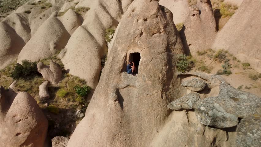 4K aerial drone pulls back and ascends over a traveler sitting in a troglodyte house in Cappadocia, Turkey. Reveals the surrounding fairy chimneys and valleys under natural sunlight. Ideal for travel,