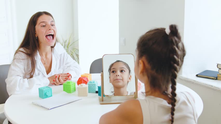 Speech therapist engages with young girl using mirror and blocks for interactive learning in cozy therapy session at bright room