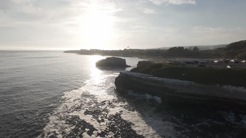 RAW ungraded sunset drone along Pacific coast sea cliffs and pocket beach — backlit surf, wave-cut platform, bluff-top park overlook, marine layer haze