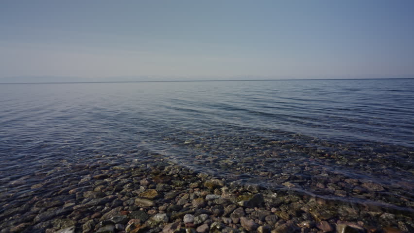 Tranquil lake surface reflecting cloudless azure sky, smooth stones gleaming through pristine waters, embodying serene natural landscape with meditative stillness, no people, nobody, slow motion.