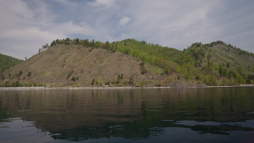 Scenic circum-Baikal railway winding along rocky Baikal lakeshore, forested slopes mirroring mountainous landscape under overcast sky. Concept of vacation and travel in Russia. Shooting in slow motion
