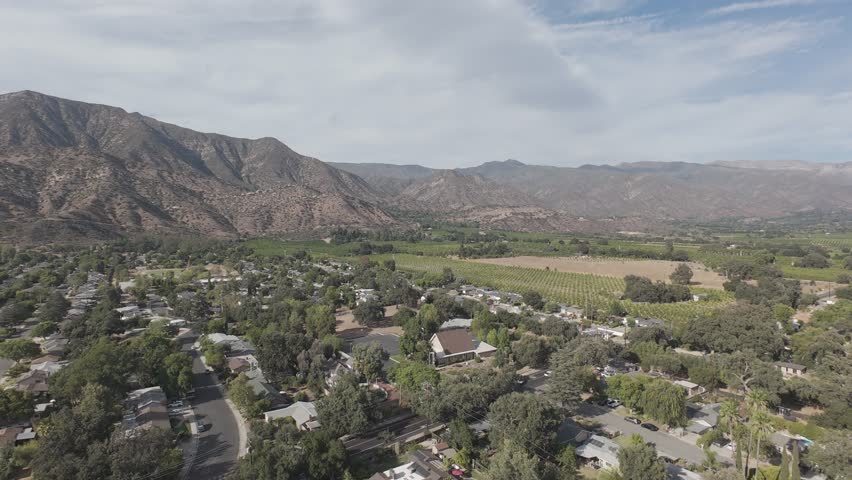 RAW ungraded drone flyover of a foothill valley town and adjacent vineyards under altostratus and stratocumulus cloud deck – urban and agricultural land use mosaic in Southern California
