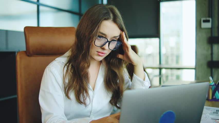 Young woman experiencing stress in a modern office environment while working on a laptop during a busy day filled with paperwork and deadlines