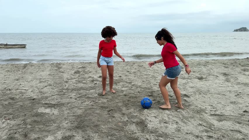 Two young girls enjoy a friendly game of beach soccer, running and kicking a blue ball on the sand