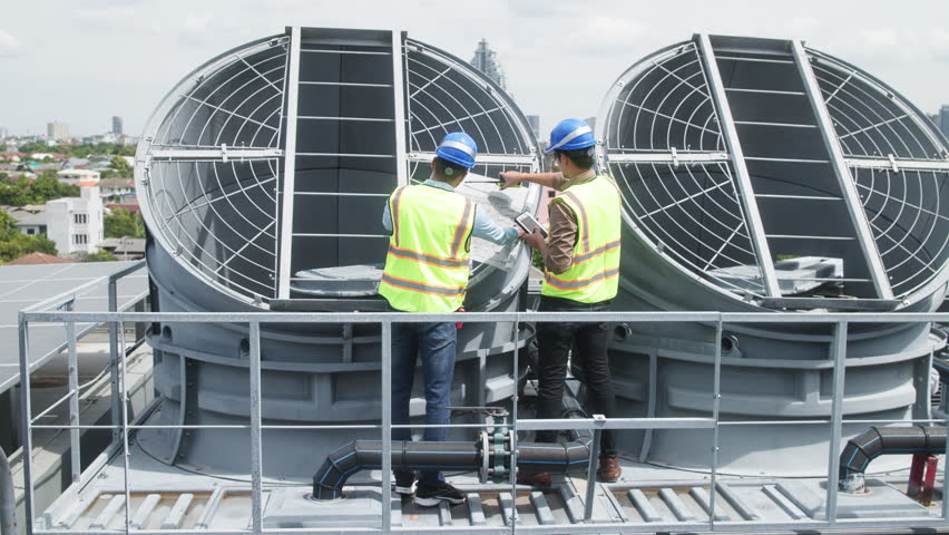 Two construction engineers in safety gear examine industrial HVAC units on rooftop, pointing and discussing project progress and setup