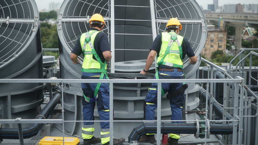 Two construction engineers in safety gear examine industrial HVAC units on rooftop, pointing and discussing project progress and setup