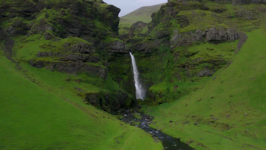 Beautiful waterfall cascades over rocky cliffs into a flowing river, surrounded by lush green forest and mountain scenery in New Zealand.