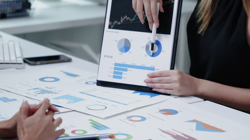 Person analyzing financial charts on a tablet beside a desktop showing candlestick graphs, reviewing reports and metrics in a modern office.