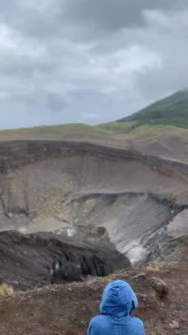 Volcanic crater of Mount Lokon in Tomohon, Indonesia, featuring a desolate and rugged terrain with a lush green peak under a dramatic, cloudy sky.
