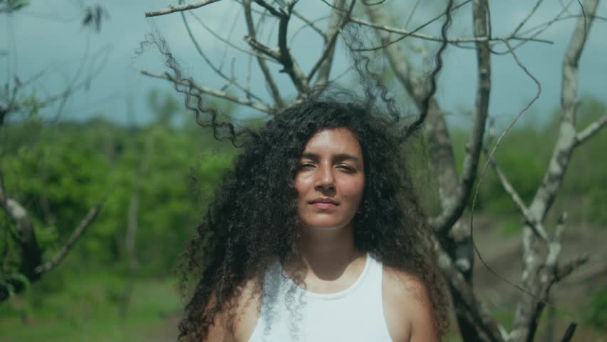 A woman stands motionless in front of a tree, her long curly hair caught in the branches as the camera pulls back.