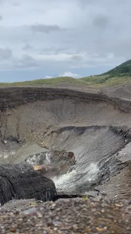 Volcanic crater of Mount Lokon in Tomohon, Indonesia, featuring a desolate and rugged terrain with a lush green peak under a dramatic, cloudy sky.