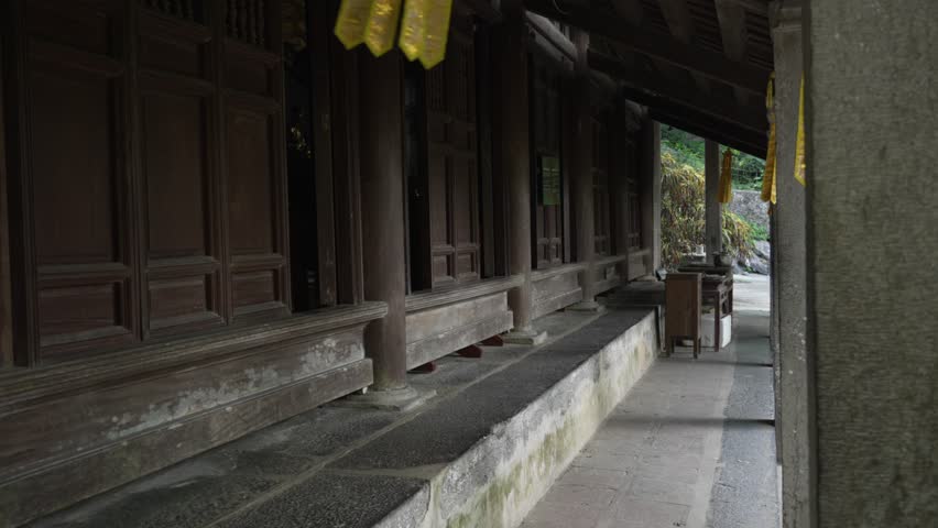 An empty wooden corridor at Bich Dong Pagoda in Ninh Binh leads past carved pillars toward soft greenery, evoking quiet solitude and temple calm, slow reveal handheld shot.