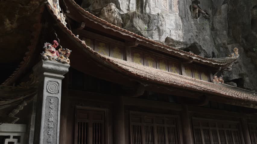 Ornate tiled roof of Bich Dong Pagoda beneath a weathered limestone cliff with carved reliefs and cave niches in Ninh Binh, Vietnam, finished with a calm upward tilt handheld shot