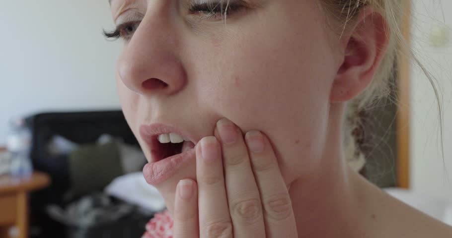 Close-up of a woman holding her cheek and reacting to tooth pain. The facial expression and hand gesture indicate discomfort, possibly from dental or gum issues.