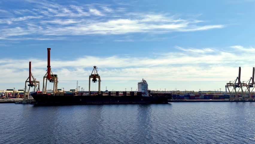 Massive cargo vessel is docked at an industrial shipping port, flanked by towering cranes and stacks of containers under a bright blue sky with scattered clouds.