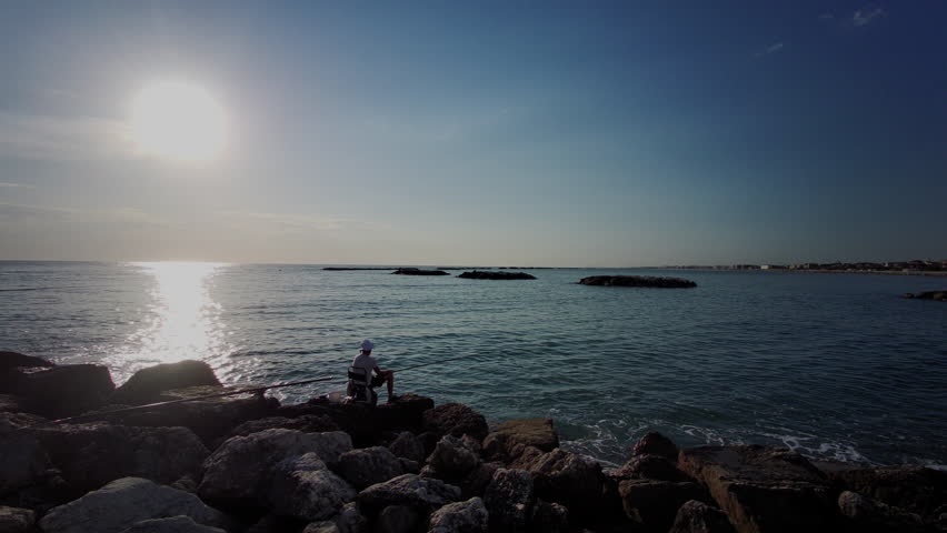 A silhouette of a person fishing from a rocky breakwater at the shoreline during sunset, with the sun