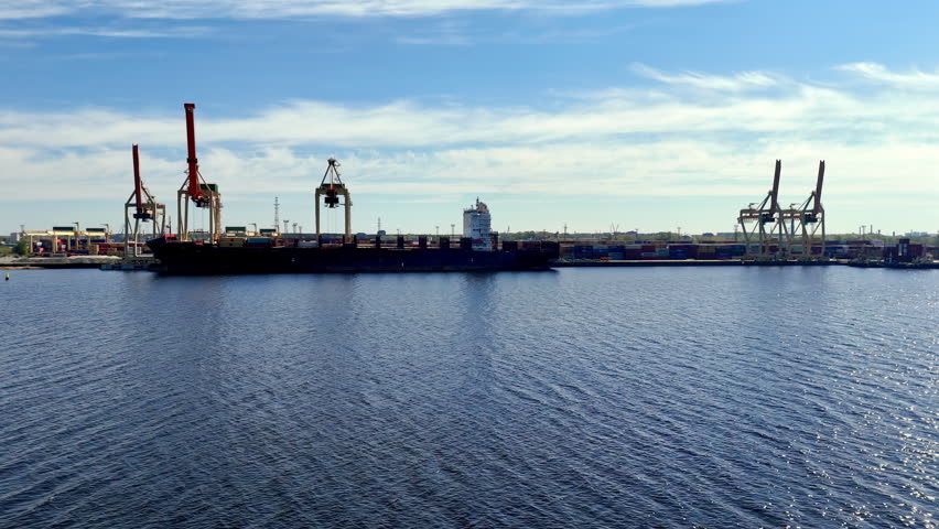 Large cargo ship rests at port terminal with cranes prepared for loading and unloading containers, set against calm harbor waters and a bright blue sky with clouds.