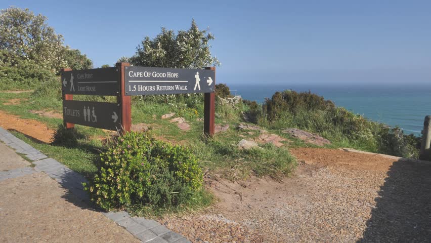 Sign board giving directions to Cape Point and Cape of Good Hope hiking trails. Grass and bushes blow in wind