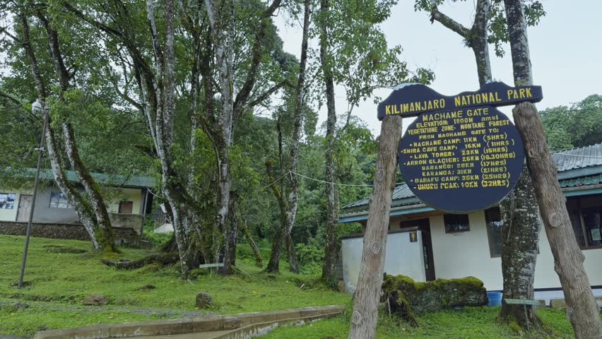 Panning shot shows a wooden information signboard at a park gate on Mount Kilimanjaro, with a small building and lush green trees in the background.